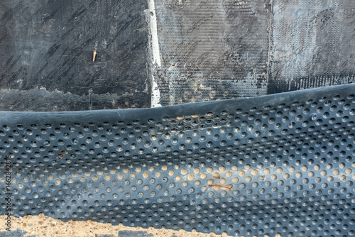 Foundation waterproofing with protective dimpled drainage membrane installed around concrete wall base, preventing moisture infiltration, improving durability, and protecting building structure