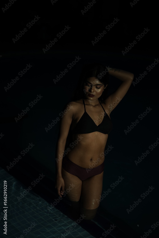 Fototapeta premium Woman Sitting by Pool at Night, Legs in Water, Bikini Swimwear