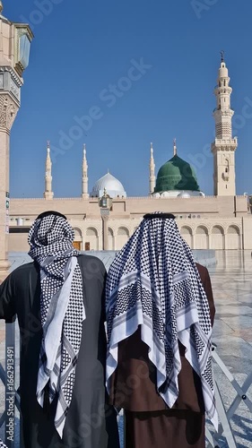 Static vertical back shot view of two unidentified muslim men wearing keffiyeh look on at the green dome at the compound of Nabawi mosque in Medina, Saudi Arabia.