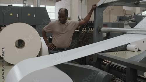 Mature Black print shop worker standing in front of paper sheet cutting machine controlling process
