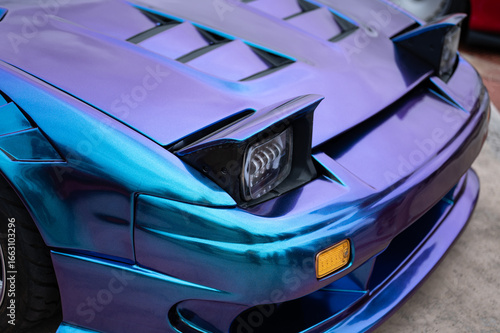 Close-up of the pop-up headlights on a classic sports car with a stunning iridescent blue and purple vinyl wrap, highlighting its JDM style.