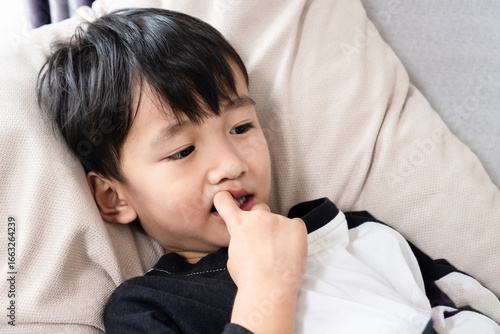 Asian boy biting his nails at home, showing signs of childhood anxiety, stress, emotional problems, and bad habits that affect kids’ health and behavior