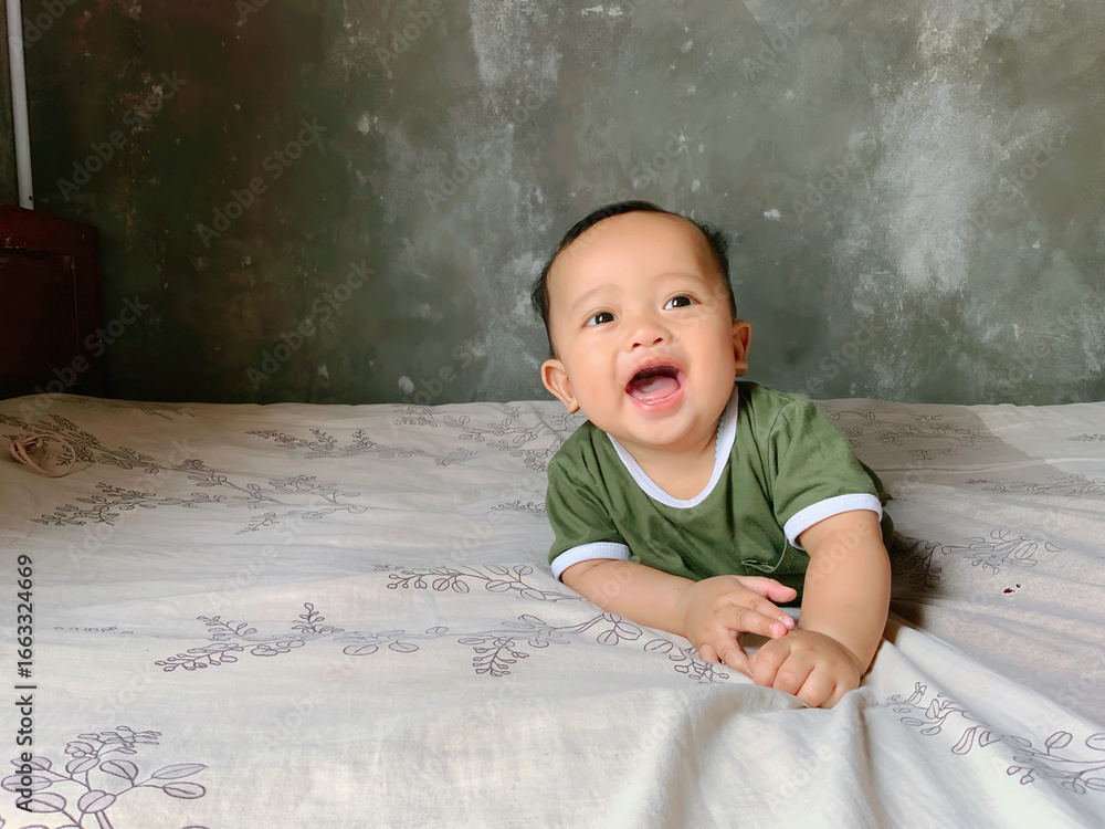 Fototapeta premium A happy baby boy is lying on his stomach, propped up on his hands, and looking directly at the camera with a wide, joyful smile. He is wearing a simple green t-shirt with white trim.