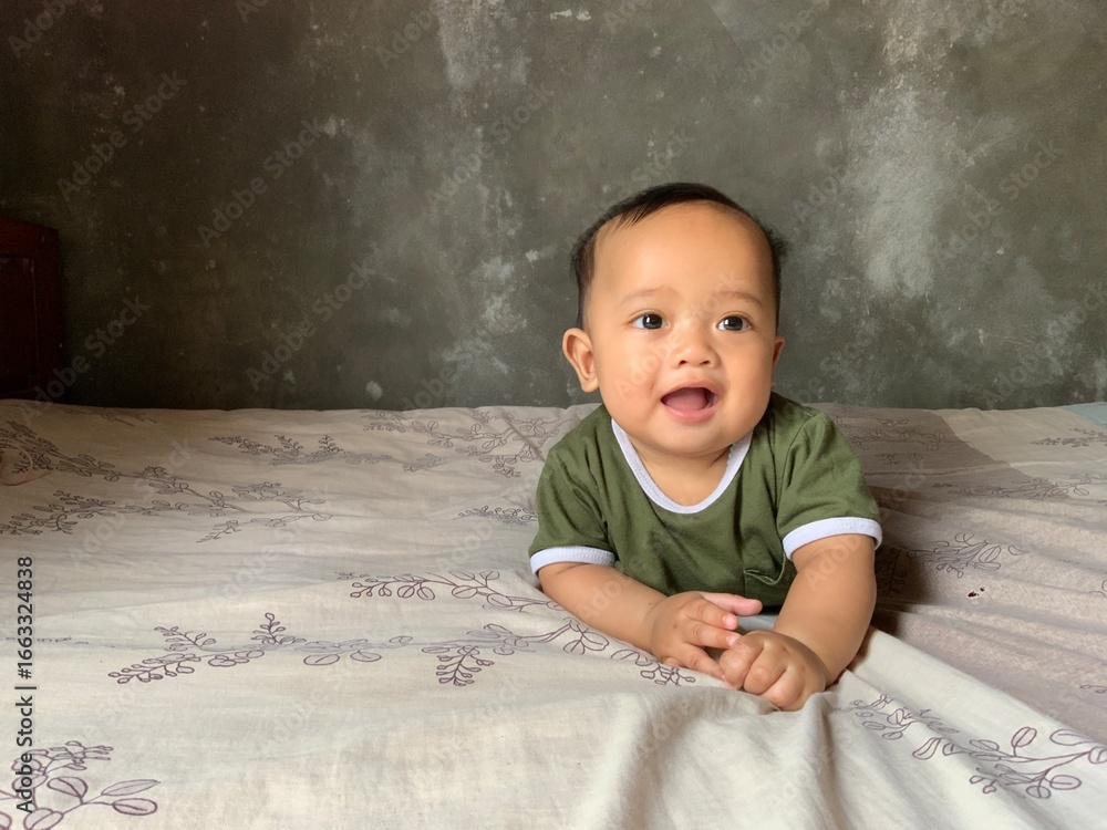 Fototapeta premium Cute baby boy lying on his stomach on the bed, smiling happily in natural light with a plain wall background.