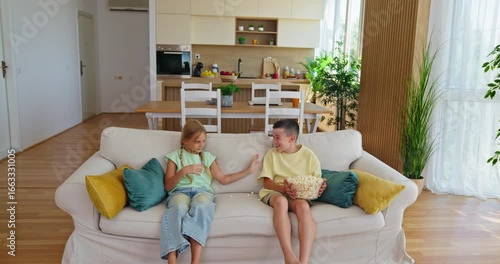 Brother and sister fight over popcorn. Brother laughing, holding bowl of popcorn while watching tv with upset sister sitting on sofa at home