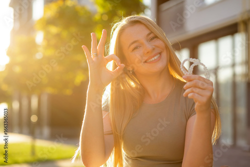 Smiling woman holding orthodontic aligners with hand gesture in sunlight
