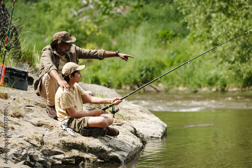 Obraz na plátně Middle aged man guiding boy fishing on rocky riverbank, man pointing toward wate