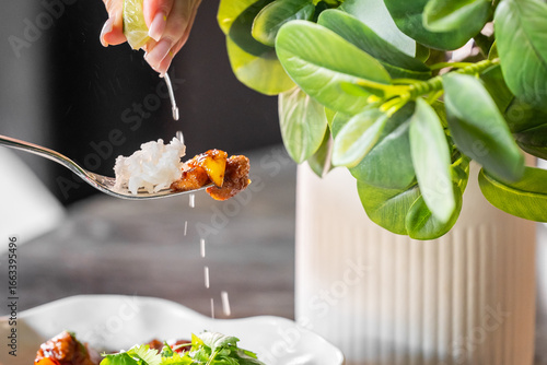 Close-up of hand squeezing lemon over spoonful of rice and meat, with juice droplets falling. Fresh garnish and green plant in background.