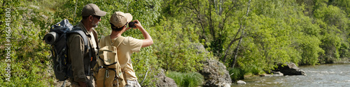 Middle aged man and boy standing on riverbank fishing together, both wearing backpacks, boy holding fishing rod, surrounded by lush green trees and flowing water