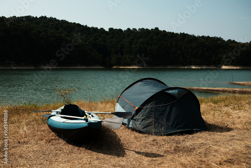 Inflatable kayak and blue camping tent on the shore of a calm lake surrounded by forested hills
