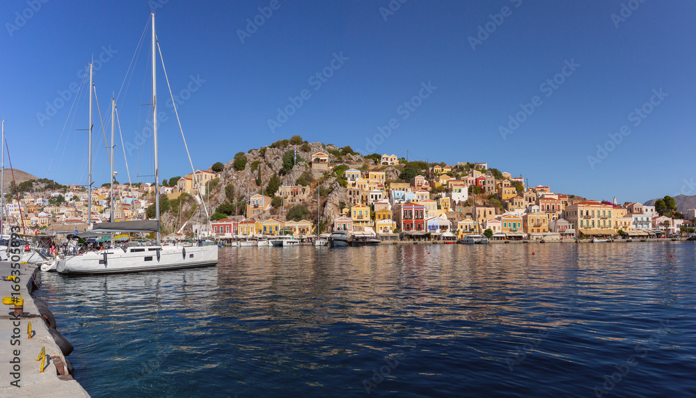 Fototapeta premium Panoramic waterfront view of Symi harbor in Greece with yachts and colorful hillside houses