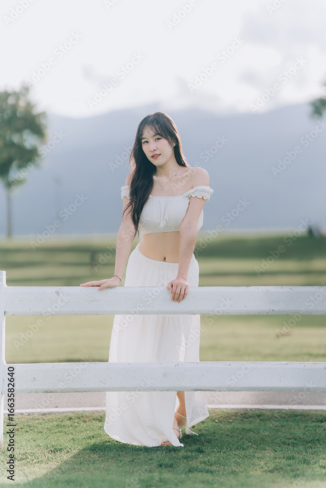 Naklejka premium Elegant Pose by Fence: A graceful woman in a flowing white dress leans against a rustic fence, her presence enhanced by a serene outdoor setting.