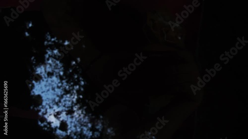 In a freshly dug grave, the gravedigger, or murderer, uses a shovel to fill in the earth, burying the dead man. Low angle. POV. Slow motion.