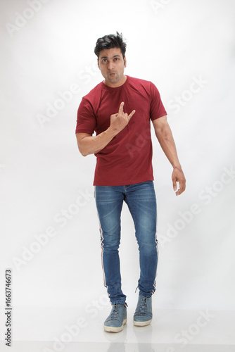 Young man wearing a red t-shirt and jeans making a rock and roll hand sign while posing playfully against a white background, reflecting modern casual fashion, energetic personality, youth lifestyle