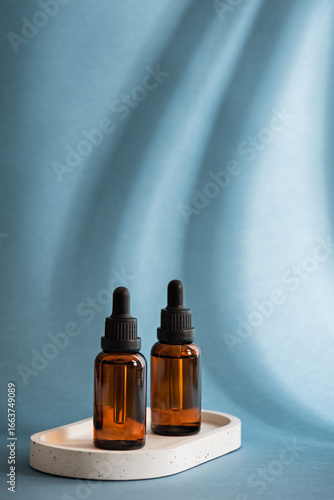 Two amber glass dropper bottles with cosmetic serum or essential oil on ceramic tray on pastel blue background with shadows, vertical