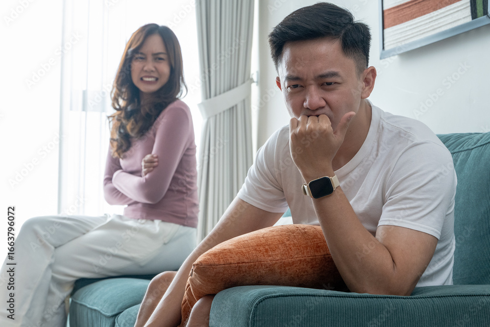 Obraz premium Young Asian couple sitting on green sofa indoors, looking upset after argument. Man in white t-shirt with smartwatch appears stressed, woman in pink top looks angry with arms crossed