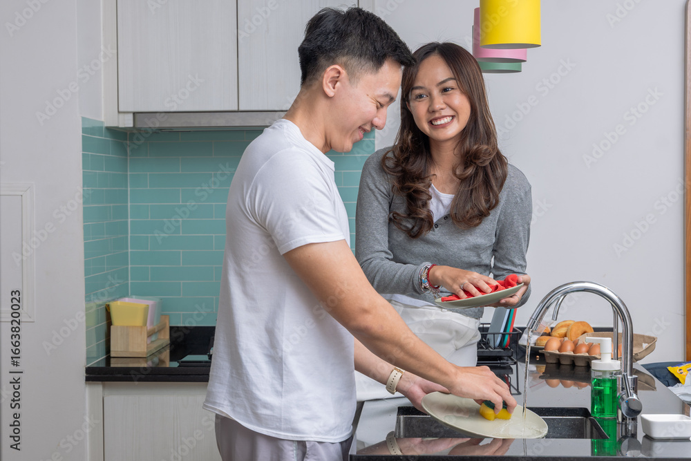 Obraz premium Smiling young Asian couple doing dishes together in modern kitchen. Man washing plates while woman dries, showing teamwork, happiness, and domestic lifestyle at home.