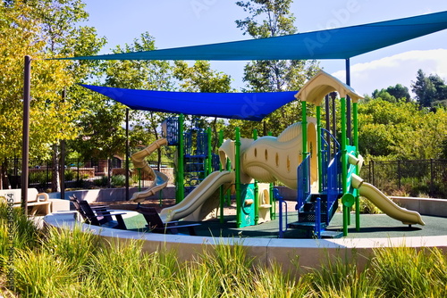 Fototapeta Naklejka Na Ścianę i Meble -  Colorful playground in Corona, California, featuring slides, shade covers, and play structures surrounded by trees in a sunny, family-friendly park setting.