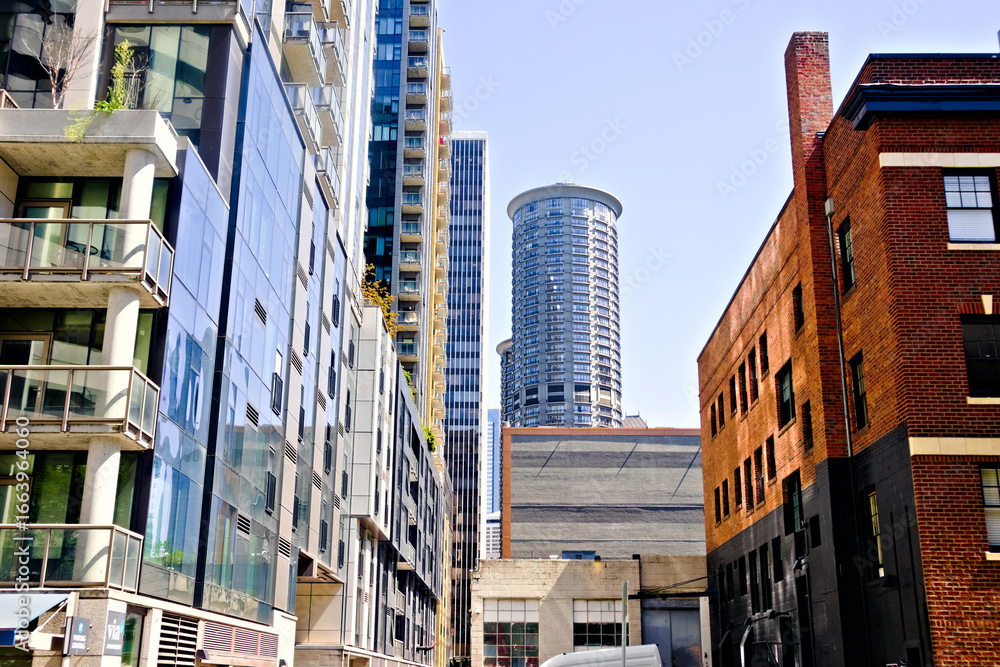 Fototapeta premium A narrow urban alley framed by brick and glass facades, leading to sleek high-rises and a round tower, blending industrial charm with modern city architecture.