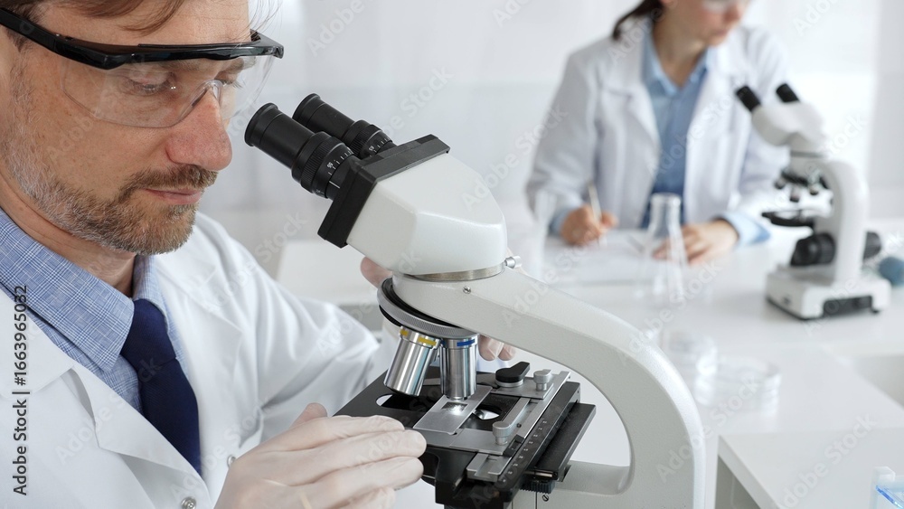 Fototapeta premium Researcher examining sample through microscope, wearing safety eyewear in sterile laboratory setting, surrounded by scientific glassware. Medicine, healthcare and science concept