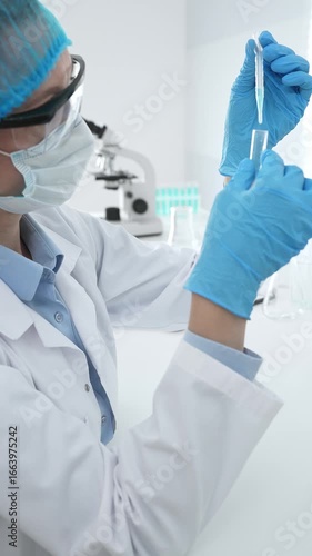 Medical science researcher wearing blue protective gear of cap, gloves and googles, is examining liquid in test tube in laboratory workspace. Medicine and health care concept