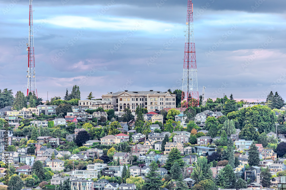 Fototapeta premium Colorful hillside neighborhood with dense houses, historic building, and tall red-white radio towers against a dramatic sky. Perfect stock image for urban landscapes, architecture, and city skylines.