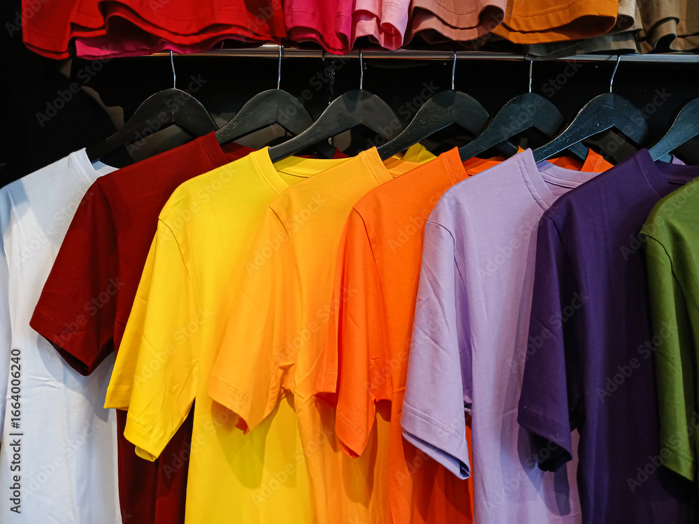 Obraz premium A variety of colorful plain t-shirts hanging on a rack. This vibrant rainbow of casual apparel is perfect for fashion, retail, and shopping concepts.