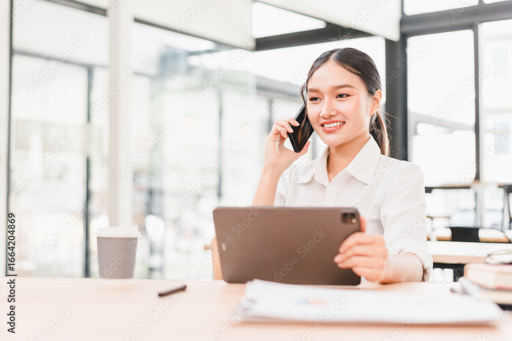 Obraz premium Young woman business attire sitting at desk holding tablet talking on smartphone smiling modern office natural light professional confident productive