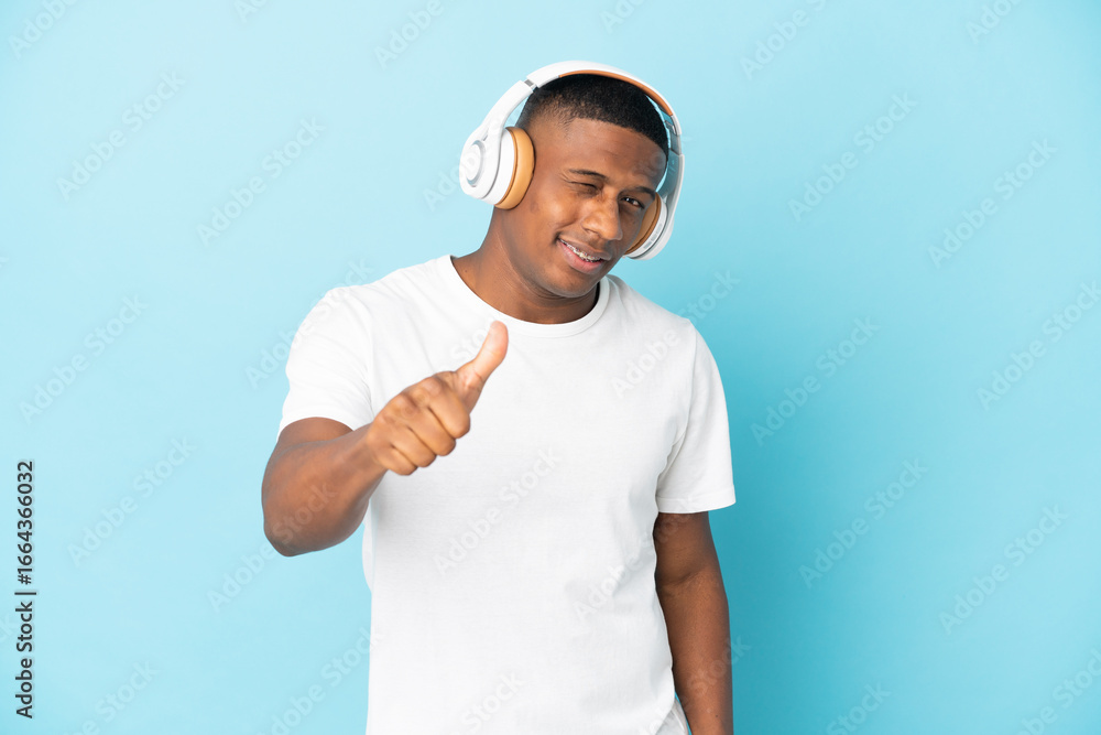 Naklejka premium Young latin man isolated on blue background listening music and with thumb up