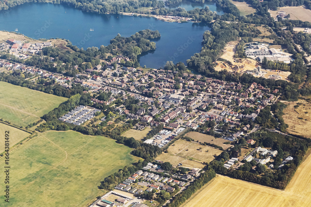 Fototapeta premium Aerial view of small town with lake. Houses and streets surrounded by green fields. Scenic countryside landscape with blue water and rural settlement. Summer day in natural environment from above.