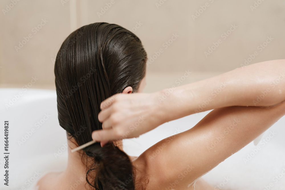 Naklejka premium Relaxed woman with long, wet hair combing her hair while taking a bath, enjoying a moment of self-care and tranquility in a serene bathroom setting.