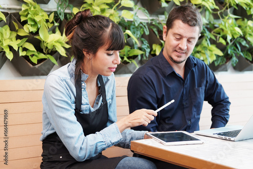 Smiling Caucasian barista couple or partnership wearing apron and consulting for marketing planning idea with tablet in the coffee shop. Start up for small cafe business owner concept.