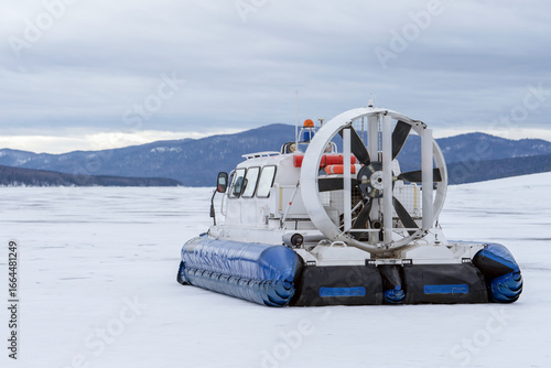 A hovercraft glides effortlessly over a snow-blanketed lake, surrounded by a serene winter landscape. The snow-covered surface reflects the cloudy sky, while distant mountains provide a picturesque