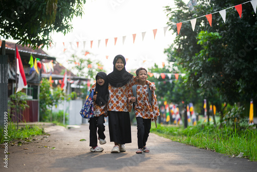 Three indonesia children happily walking together on a village road, wearing matching batik clothes and carrying school bags. Joyful friendship and family moment.