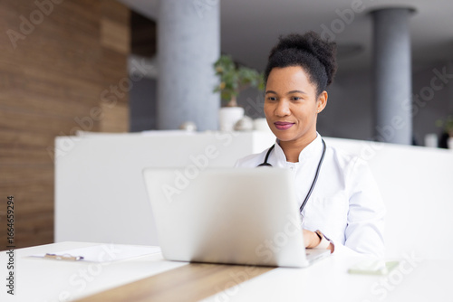 Professional female physician typing on laptop during telehealth consultation, accessing patient records in contemporary medical setting