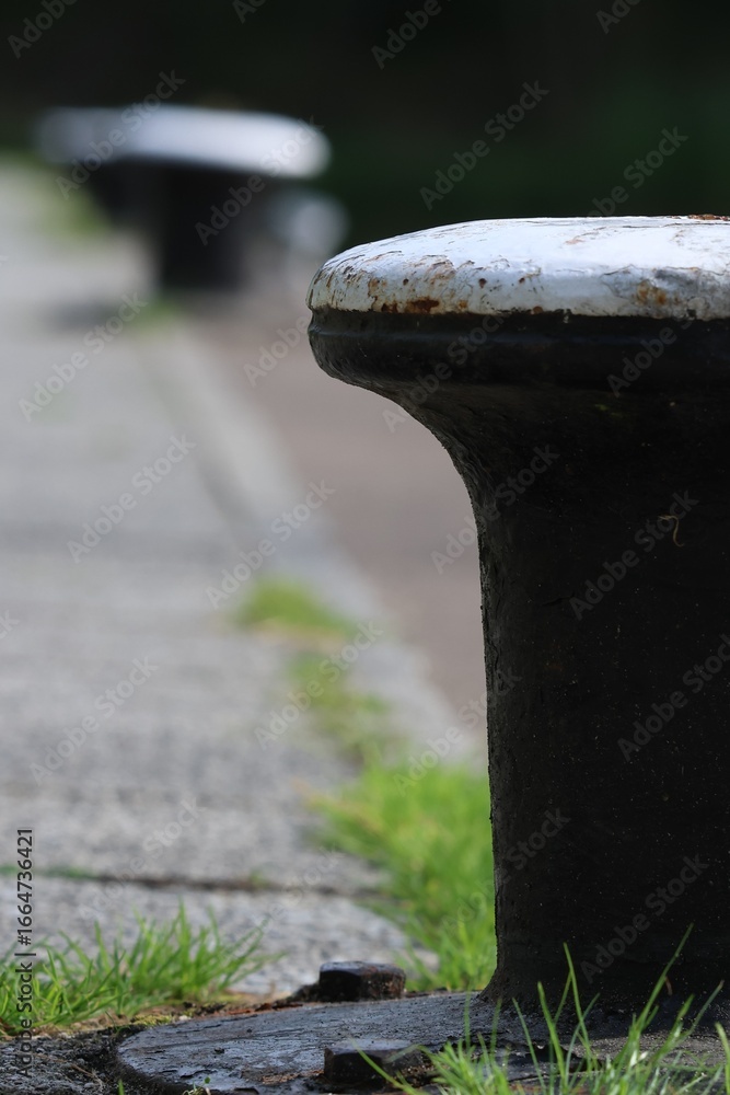 Φωτογραφία Close-up of a weathered mooring bollard on a quiet riverside dock, surrounded by lush park greenery, evoking calm and peaceful waterside ambiance