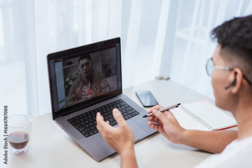 Fototapeta premium Asian man in glasses and earbuds gesturing hands while speaking on laptop video call at bright home office desk. Suitable for remote meetings, webinars, and online collaboration lifestyle themes