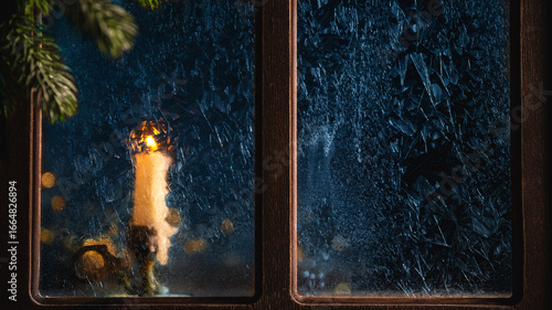 Single white candle is burning behind a frozen window during a cold winter night, creating a warm and cozy atmosphere with the fir tree branch in the foreground