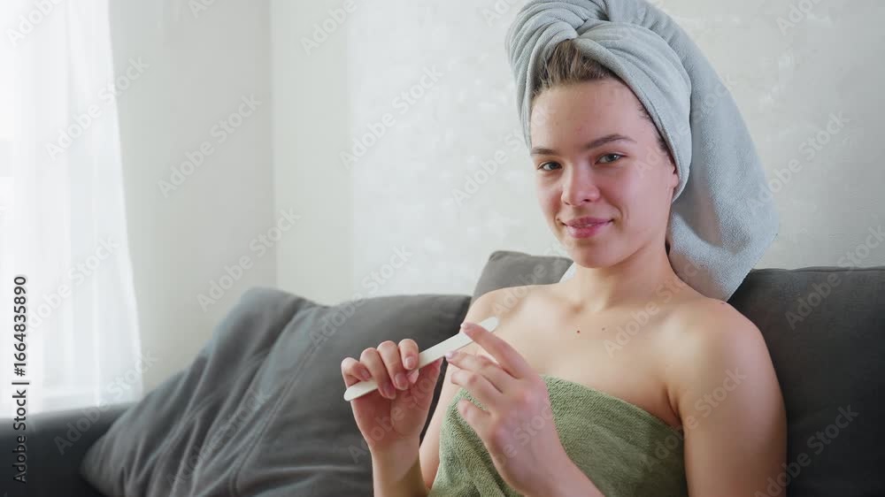 Young woman sits comfortably on couch, smiling and focused while filing nails. Wrapped in towel around chest, she exudes calm and confidence in cozy indoor setting, highlighting self-care Young woman sits comfortably on couch, smiling and focused while filing nails. Wrapped in towel around chest, she exudes calm and confidence in cozy indoor setting, highlighting self-care