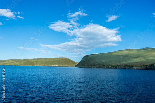 The coastal panorama of Lake Baikal under a bright blue sky, with calm water and gentle hills.