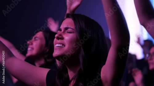 A woman with dark hair and closed eyes raises her hands in a dimly lit room with other people around