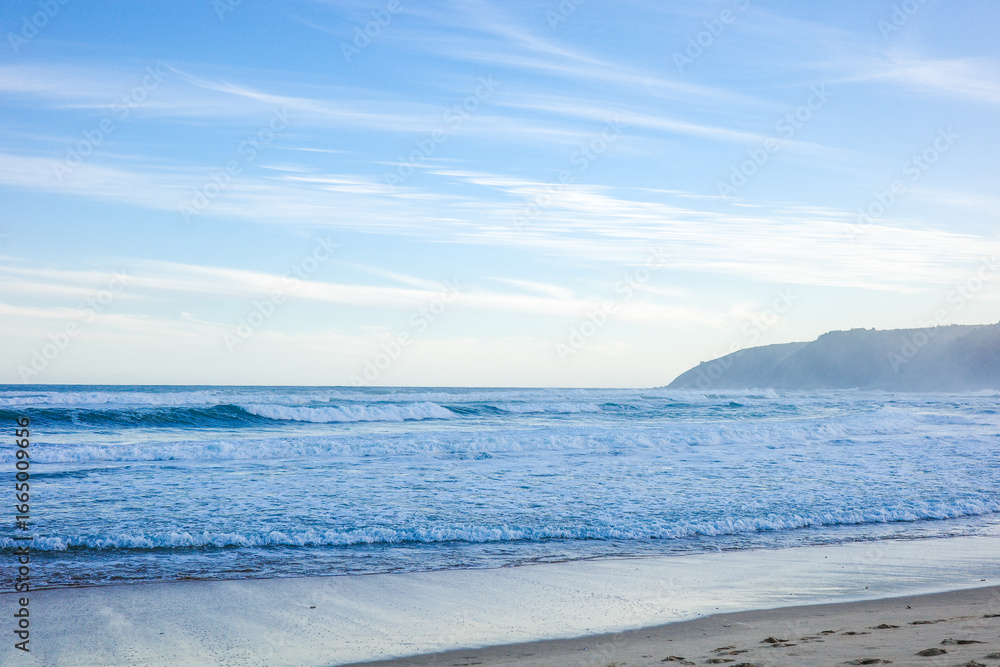 Obraz premium A calm seascape featuring gentle ocean waves rolling onto the sandy shoreline under a clear blue sky with wispy clouds and a misty coastal hill in the distance. The natural beauty of coastline
