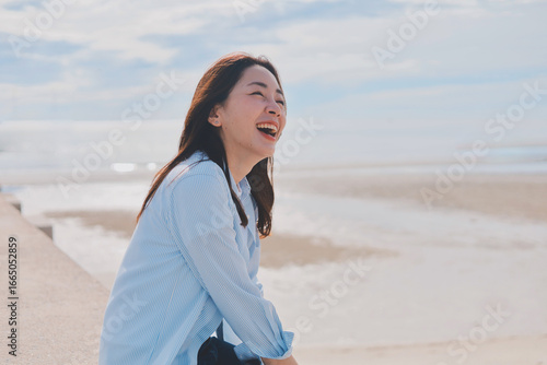 A cheerful woman wearing a casual shirt and jeans sitting in front of the blue sky background with copy space.