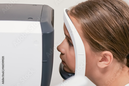 Child placing chin on a slit lamp or autorefractor during an eye examination at an ophthalmology clinic. Advanced vision testing equipment in use.