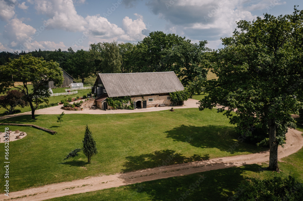 Obraz premium A large rustic countryside barn with a gray roof surrounded by a green lawn, gravel paths, trees, and a peaceful garden on a sunny day with scattered clouds.