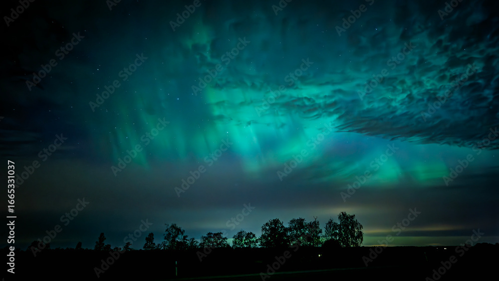 Naklejka premium Northern lights dancing behinde the dark clouds over country landscape
