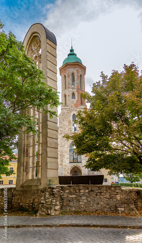 Photography Vertical shot of recreated Gothic window at Mary Magdalene Church foundation with bell tower against blue sky, framed by autumn foliage in Buda Old Town
