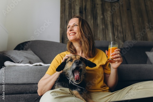 Woman relaxing at home with her dog and a glass of fresh juice. Concept of remote work, healthy lifestyle, pet companionship, and cozy home atmosphere