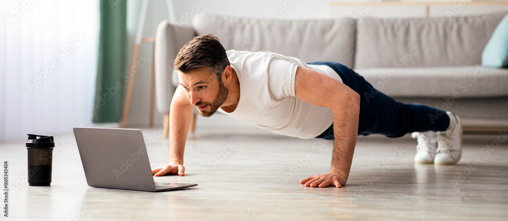 Fototapeta premium Handsome middle-aged man in sportswear doing push-ups, using laptop, having online fitness class from home, living room interior, copy space. Sporty bearded man enjoying daily training, watching video