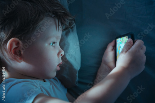 Close-up of small boy lying in bed at night, eyes wide while watching cartoons on smartphone or playing mobile video games. Screen addiction and gadget overuse. 
